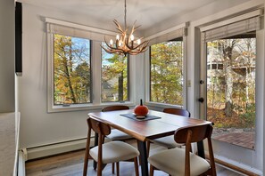 Un coin repas avec une table et des chaises en bois, un lustre et de grandes fenêtres offrant une vue sur le feuillage automnal.