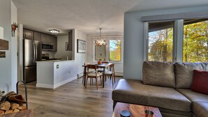 Et moderne køkken med stålemmer, et spiseområde med træstole og en lædersofa med udsigt til træer gennem store vinduer.