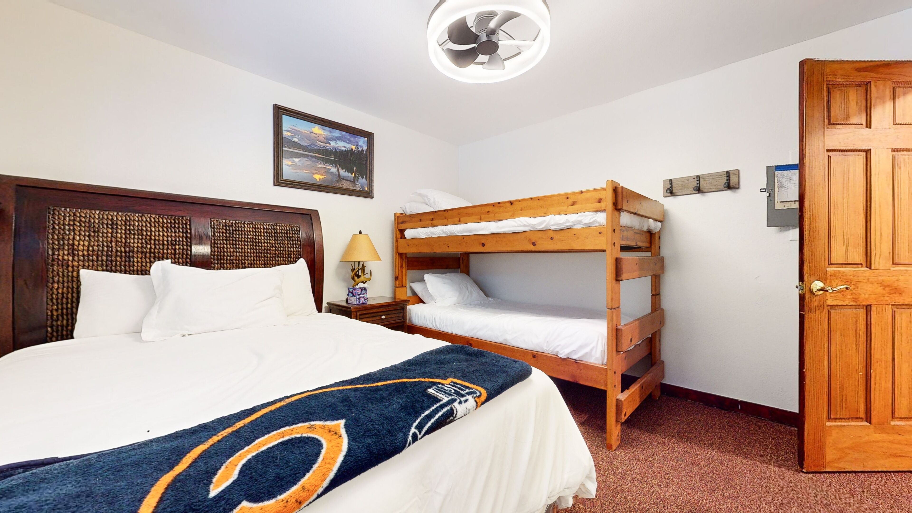 A hotel room with a large bed, a smaller bunk bed, a wooden door, a ceiling fan, and a framed picture on the wall.