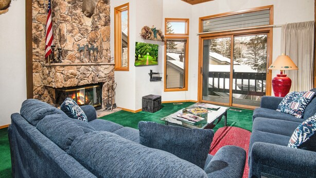 A cozy living room with a stone fireplace, a large window with a view of a snowy deck, and a comfortable blue sofa set. - Beaver Creek Townhome w/ Amenities | RP187 Stays limited to a max of 14 nites (Beaver Creek)