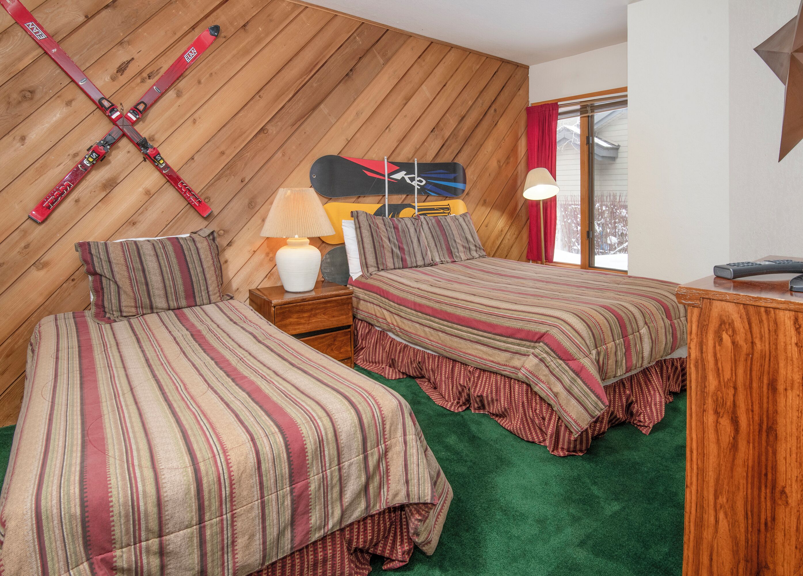 A room with two beds, a wooden wall, a lamp, and skis hanging on the wall.