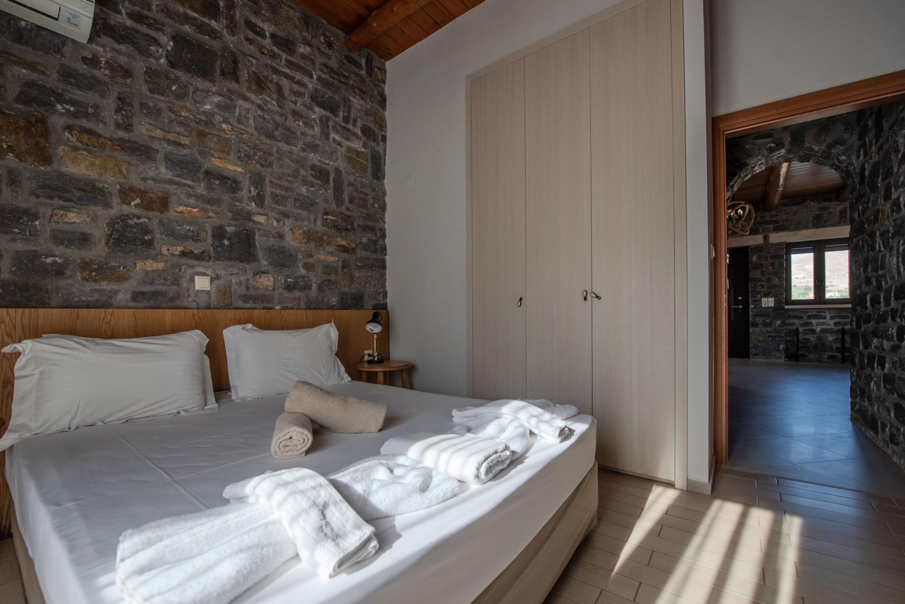 Quarto com parede de pedra, cabeceira de madeira, cama com lençóis e toalhas brancos, mesa de cabeceira com luminária e guarda-roupa de madeira.