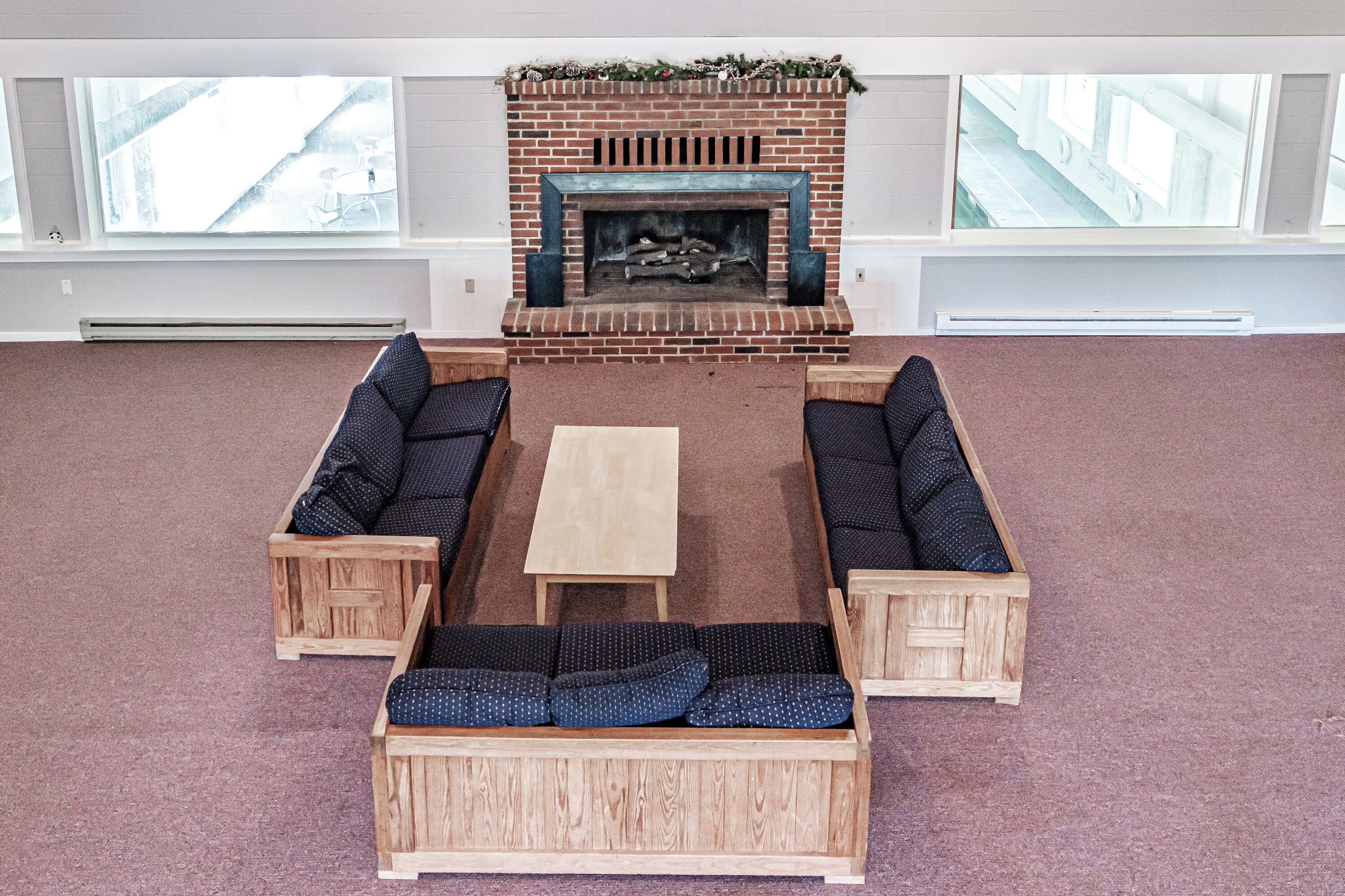A spacious room with a brick fireplace, two wooden sofas with blue cushions, a wooden coffee table, and large windows providing a view of the outside.