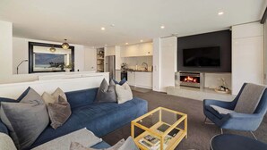 A modern living room with a blue sofa, a glass coffee table, and a flat-screen TV mounted above a fireplace. - Central Lakeview Apartment | Walk to Town | Spa (Queenstown)