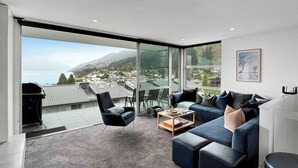A modern living room with a large sliding glass door leading to a balcony, a blue sofa set, a coffee table, and a view of a town and mountains. - Central Lakeview Apartment | Walk to Town | Spa (Queenstown)