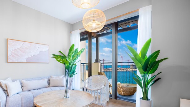 A modern living room with a round wooden table, a light grey sofa, and two potted plants. - LUX  La Mer Infinity View-Luxury Boho 1 (Dubai)