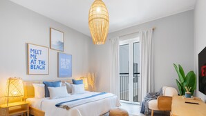 A modern bedroom with a bed, bedside lamps, a large window with curtains, a wall-mounted TV, and framed artwork. - LUX  La Mer Infinity View-Luxury Boho 1 (Dubai)