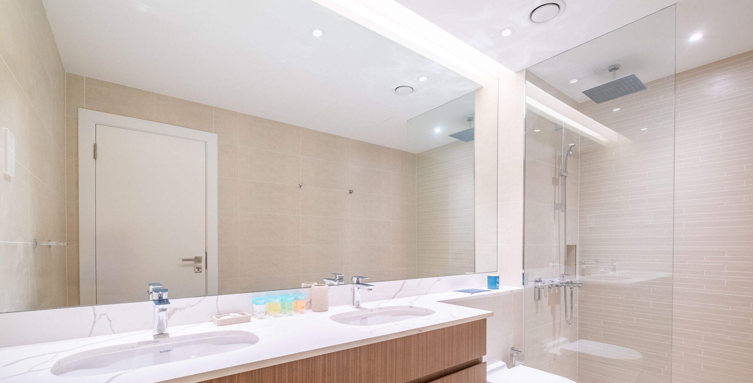 Un baño moderno con un espejo grande, dos lavamanos y una cabina de ducha de vidrio.
