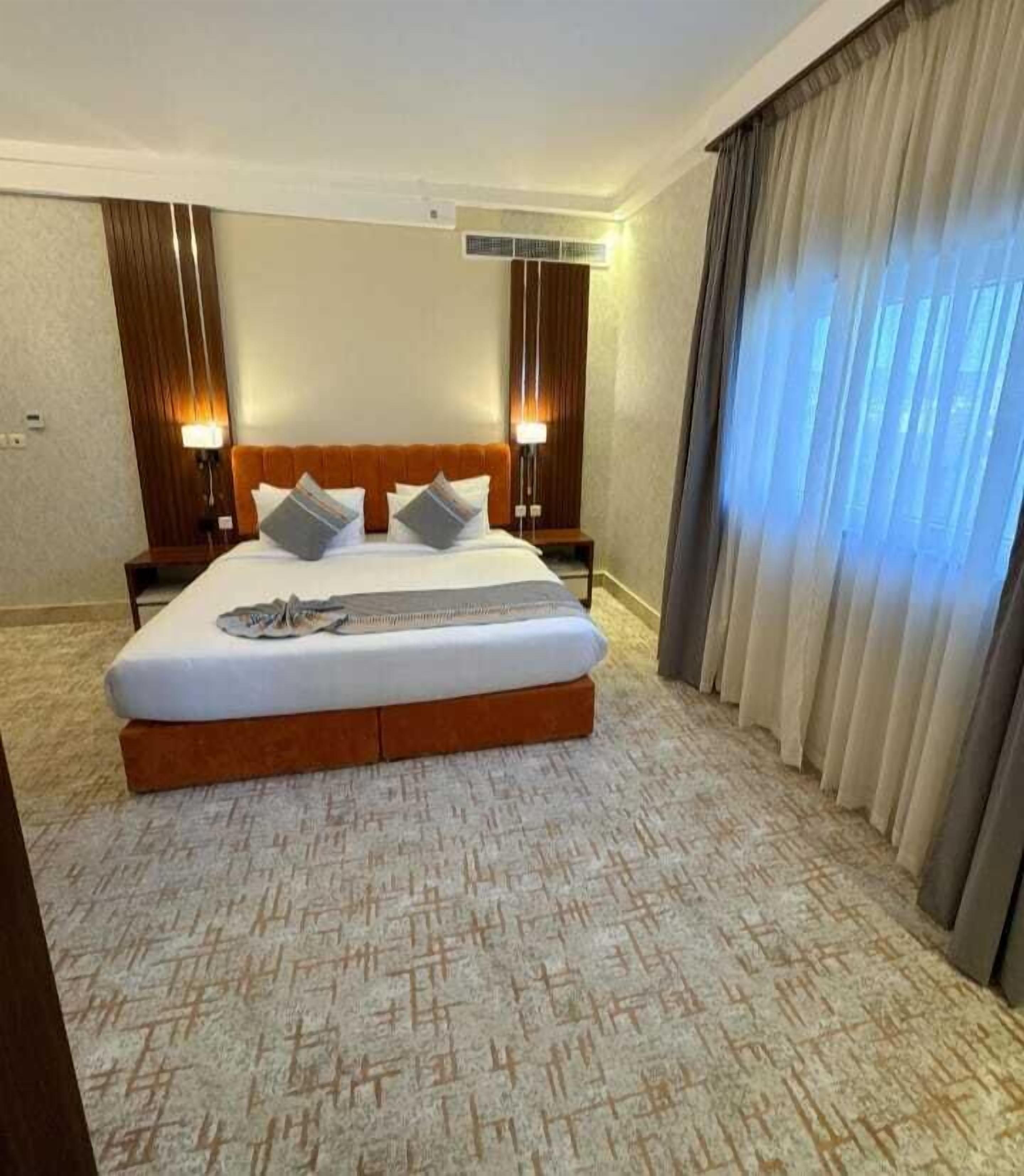 A hotel room with a large bed, two bedside tables, a headboard, and a window with curtains.