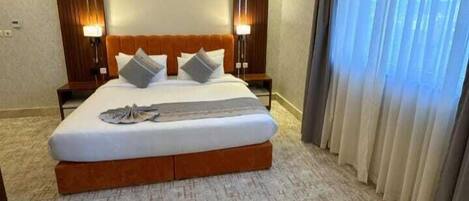 Quarto de hotel com uma cama grande, duas mesas de cabeceira, um encosto e uma janela com cortinas.