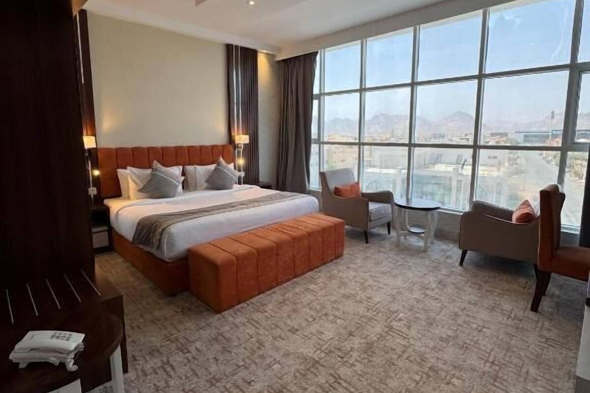 A hotel room with a large bed, a seating area with chairs, a view of the city through large windows, and a carpeted floor.