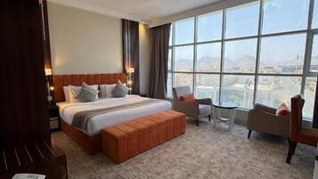 A hotel room with a large bed, a seating area with chairs, a view of the city through large windows, and a carpeted floor.
