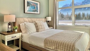 Un dormitorio con cama, dos mesitas de noche, una lámpara de noche, un cuadro enmarcado y una ventana con vistas a montañas y árboles.