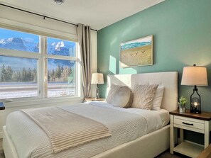 A bedroom with a large bed, two bedside tables with lamps, a window with a view of mountains, and a painting on the wall. - Brand New - Mt Rundle Views 3 Bd Townhome (Harvie Heights)