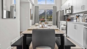 Una cocina moderna con armarios blancos, un comedor con una mesa lista para dos personas y vistas a montañas a través de un amplio ventanal.