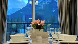 Una mesa de comedor dispuesta con platos, tazones y copas de vino, con un florero en el centro. Una ventana muestra un paisaje montañoso.