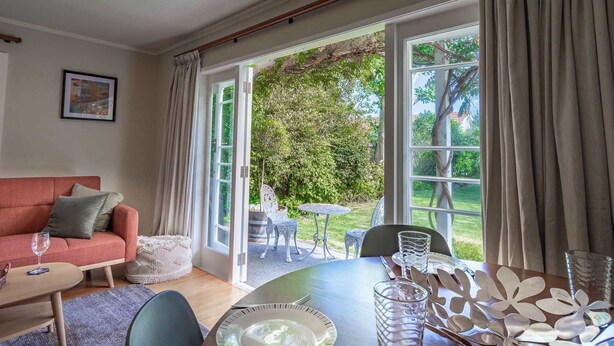 Een gezellige woonkamer met een bank, een eettafel gedekt voor twee personen en uitzicht op een weelderige tuin door grote glazen schuifdeuren.