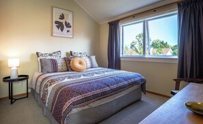 A bedroom with a bed, a bedside table with a lamp, a window with curtains, and a wall with a framed artwork.