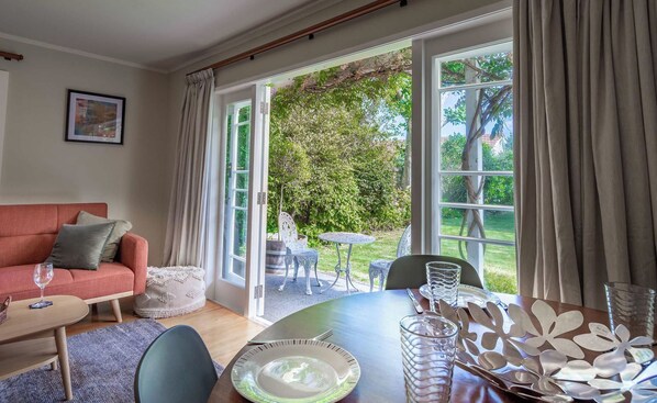 A cozy living room with a sofa, a dining table set for two, and a view of a lush garden through large sliding glass doors.