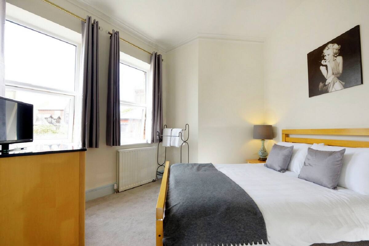 A hotel room with a bed, a television on a stand, a radiator, and a window with curtains.