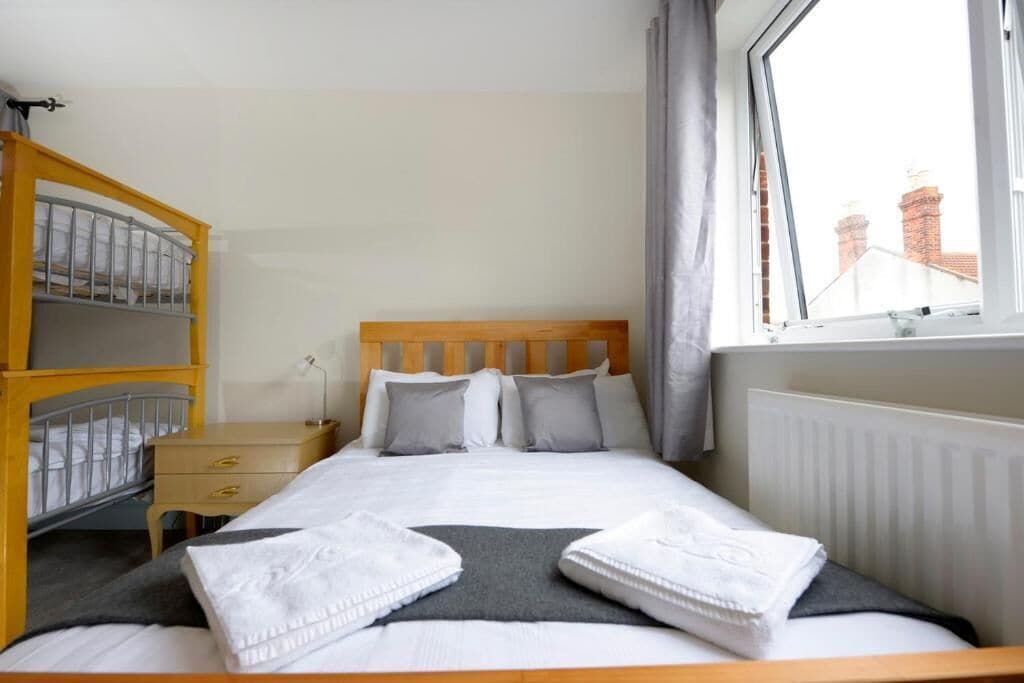 A hotel room with a bed, a wooden headboard, a nightstand, a shelf with toiletries, and a window with a view of buildings.