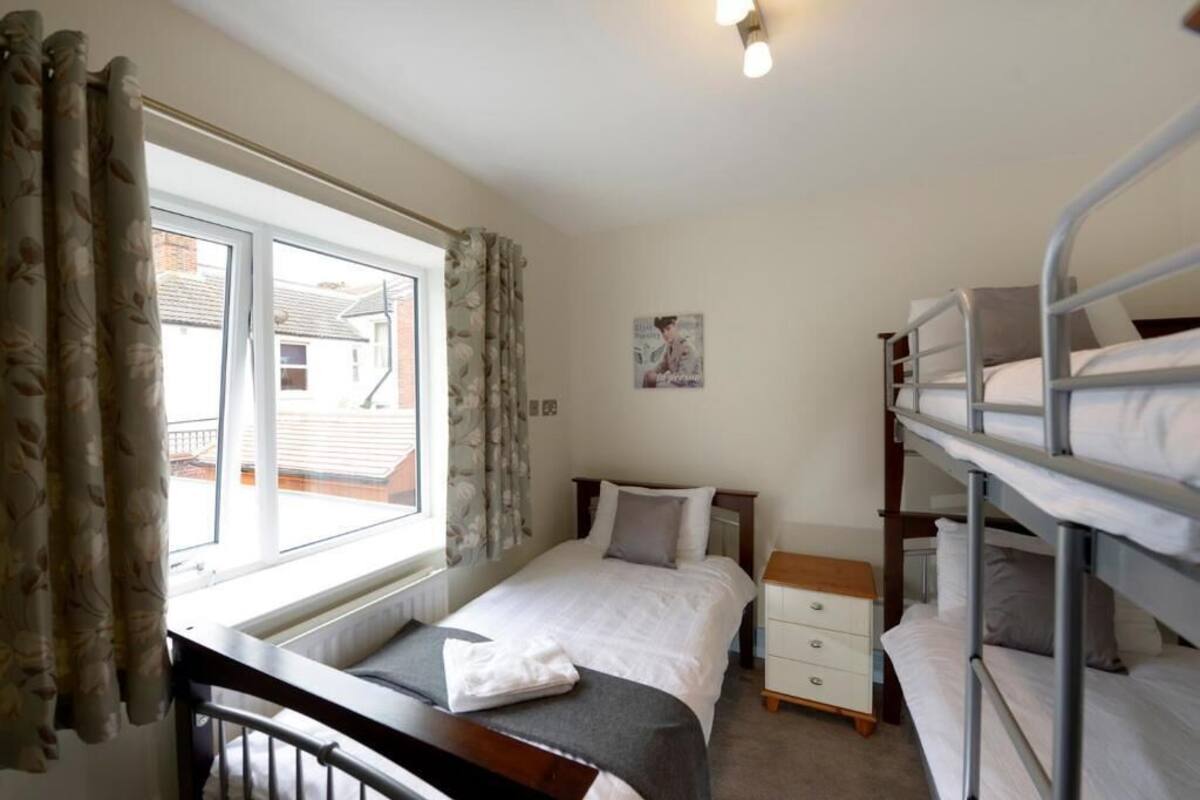 A dormitory room with bunk beds, a single bed, a window with curtains, a bedside table, and a wall-mounted light fixture.