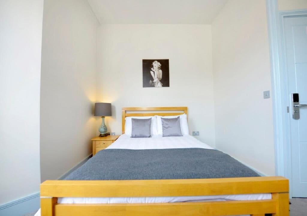 Una cama individual con estructura de madera y una manta gris, una mesita de noche con una lámpara y un cuadro en la pared.