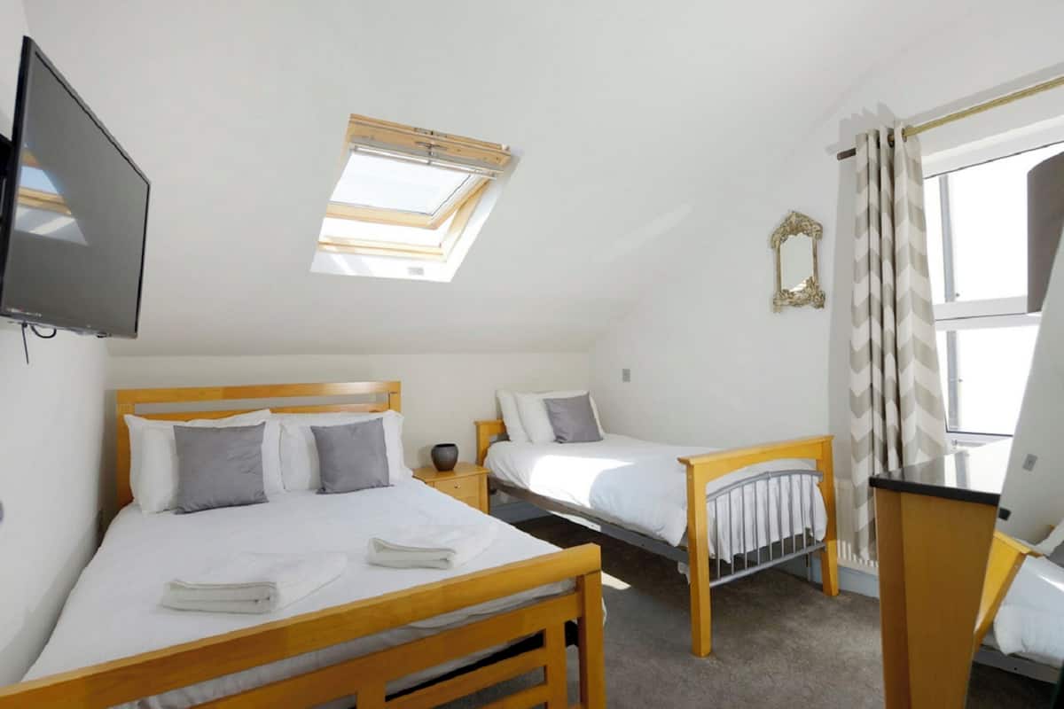 A bedroom with two beds, a TV, a skylight, a window with curtains, and a small table.