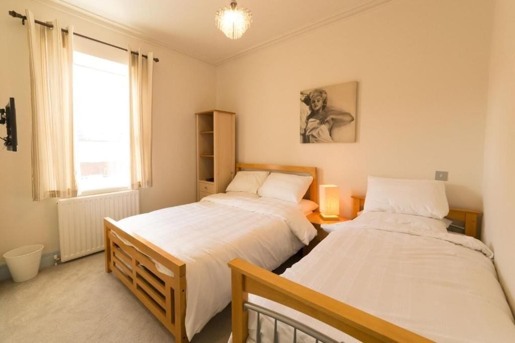 A bedroom with two single beds, a radiator, a window with curtains, a bedside lamp, and a shelf.