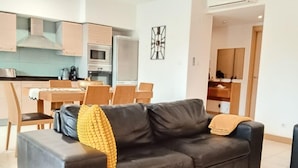 Un soggiorno moderno con un divano in pelle scura, un'area pranzo con sedie in legno, una cucina con forno a microonde e piano cottura, e un corridoio che conduce a un'altra stanza.