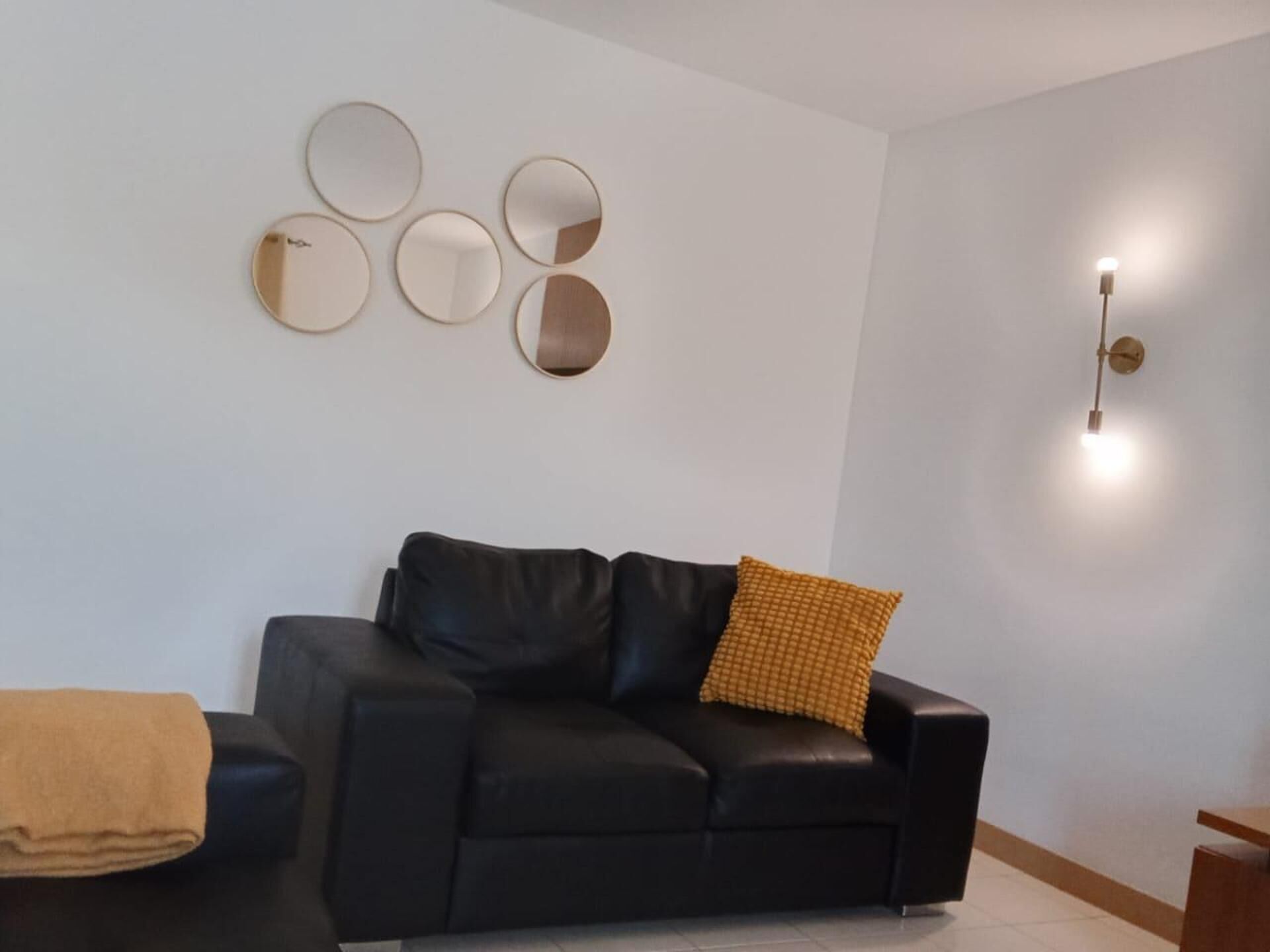 Een woonkamer met een zwarte leren bank, een mosterdgele plaid en een wandlamp.