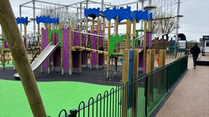A playground with various play structures, a fenced area, and a person walking in the background. - Highfield Grange Holiday Park (Clacton-on-Sea)