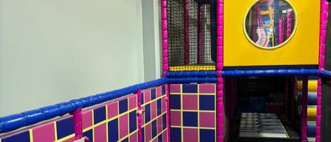 Un espace de jeux intérieur coloré, doté d’une zone d’escalade avec des filets et d’une fenêtre circulaire permettant d’observer l’espace.
