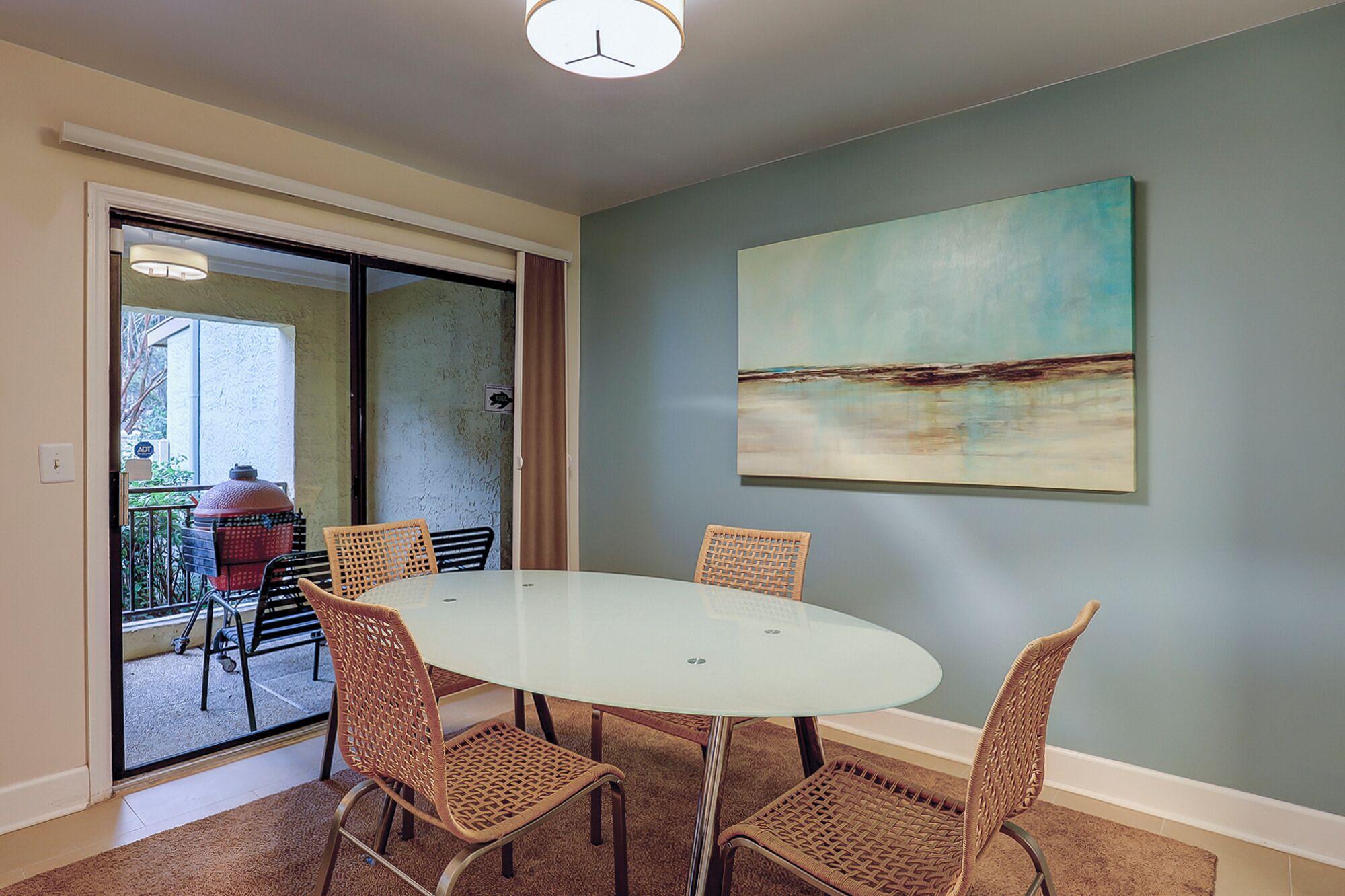 A dining area with a round table and wicker chairs, a painting on the wall, and a view through a glass door.