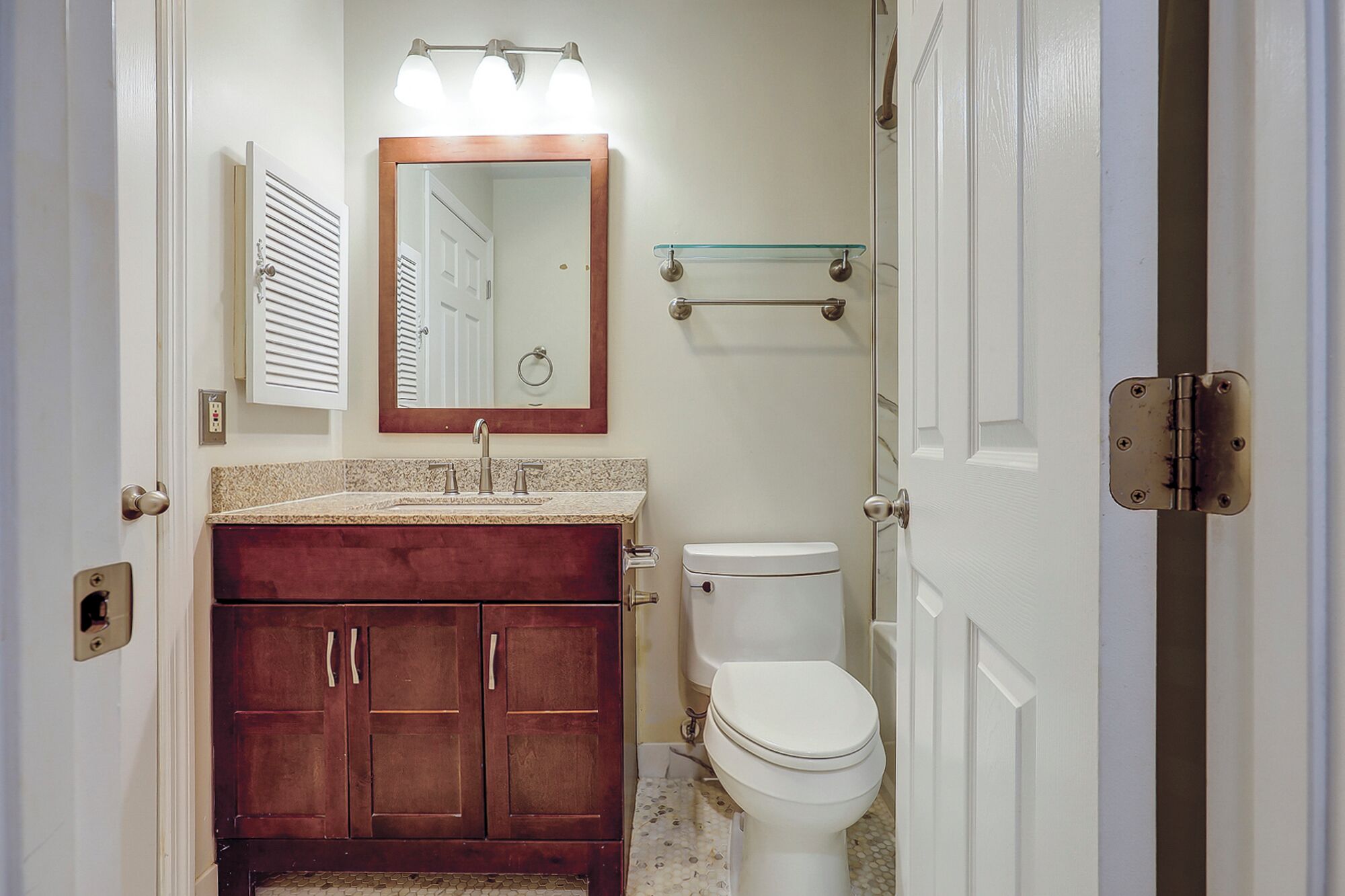 A bathroom with a wooden vanity, a white toilet, a glass shelf above the sink, and a mirror with lights.