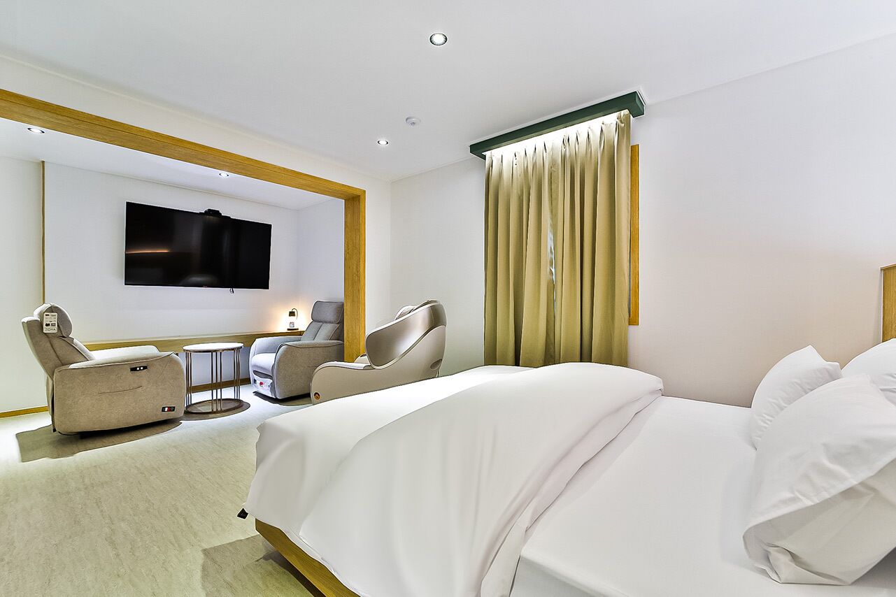 A hotel room with a large bed, two massage chairs, a small table, and a TV on the wall.