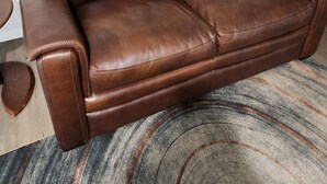 A brown leather sofa in a room with a patterned rug, a small black table, and a painting on the wall. - Avocado Executive Suite 1 (Winnipeg)
