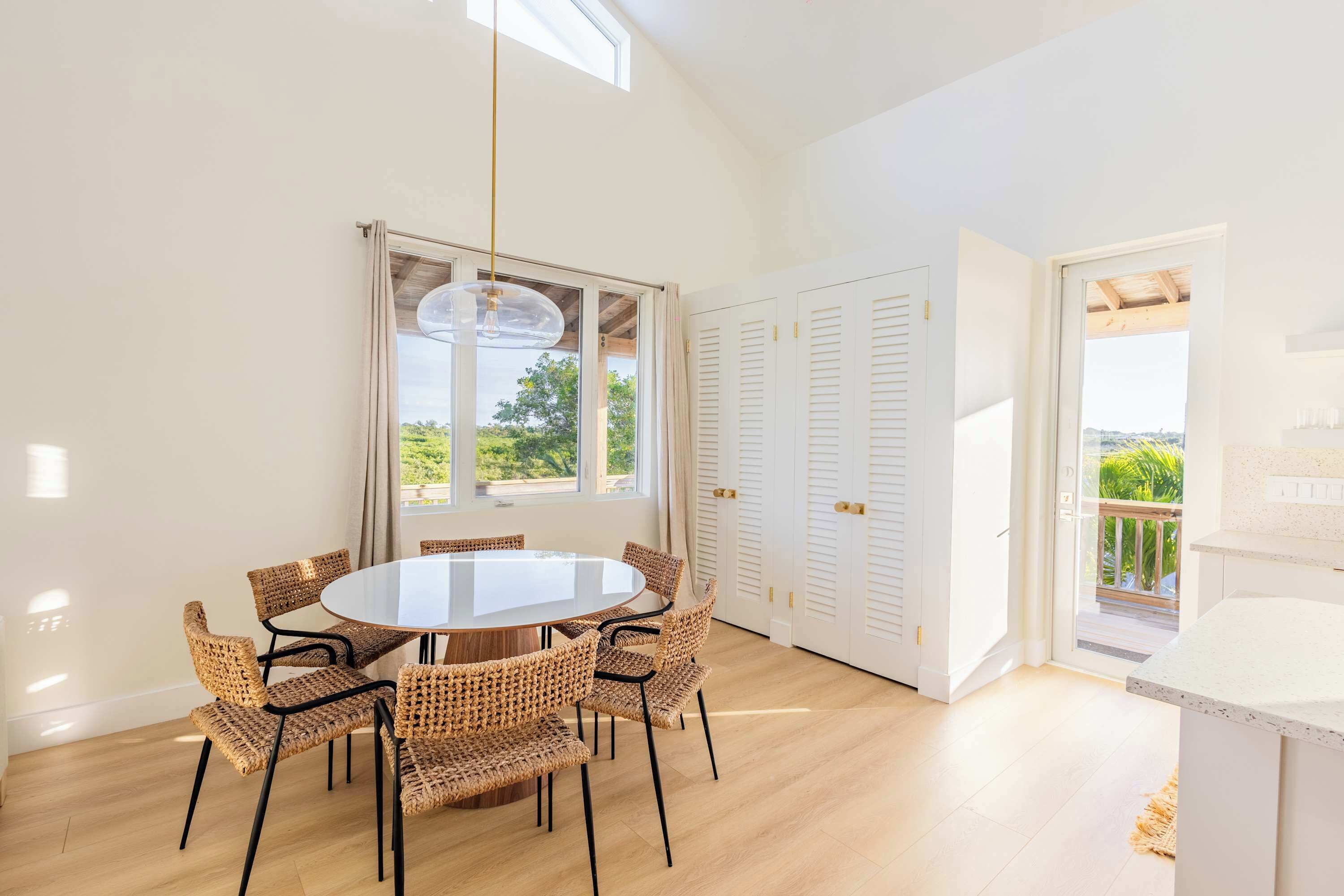 A dining area with a round table and wicker chairs, a large window with curtains, and a view of greenery outside.