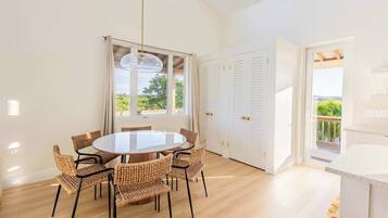 A dining area with a round table and wicker chairs, a large window with curtains, and a view of greenery outside.