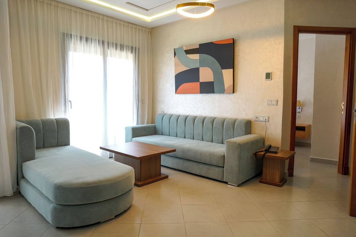 Een moderne woonkamer met een bank, een voetenbankje en een salontafel, een schilderij aan de muur en een deur die leidt naar een andere kamer.