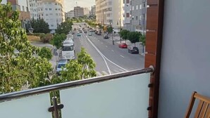 Un balcon donnant sur une rue de la ville, une petite table en bois avec un chapeau et une bouteille, et une chaise en bois.