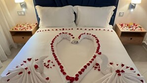 Un lit décoré de pétales de roses et de serviettes disposées en forme de cœur, avec une literie blanche et deux lampes de chevet.