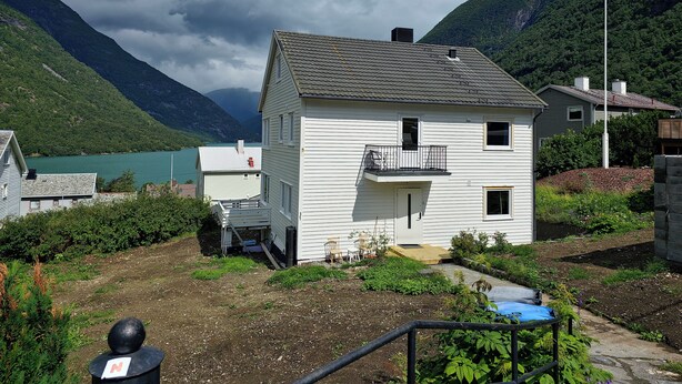 Une maison blanche à deux étages, dotée d’un balcon, entourée d’une végétation luxuriante et avec des montagnes en arrière-plan.
