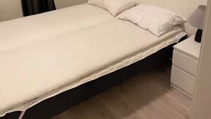 Une petite chambre avec un lit simple, un plancher en bois, des murs blancs, une fenêtre avec des rideaux et une table de chevet avec une lampe.