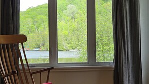 Una habitación con una mecedora, vista a un lago rodeado de vegetación y una ventana con cortinas.