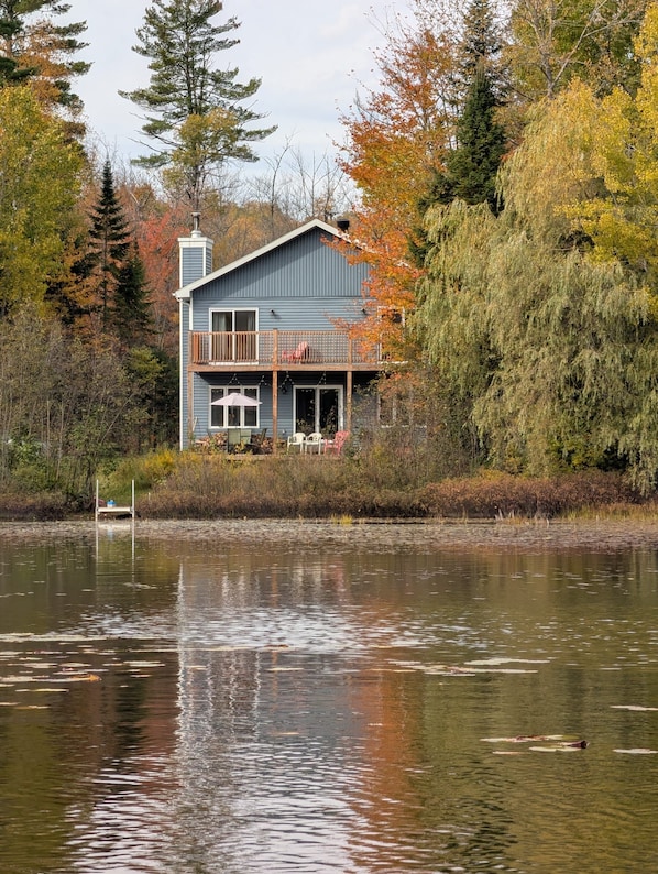 Une maison à deux étages avec un balcon, entourée d’arbres et bordée par un plan d’eau.