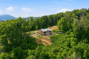A two-story house with a large deck, surrounded by dense forest and mountains in the background.