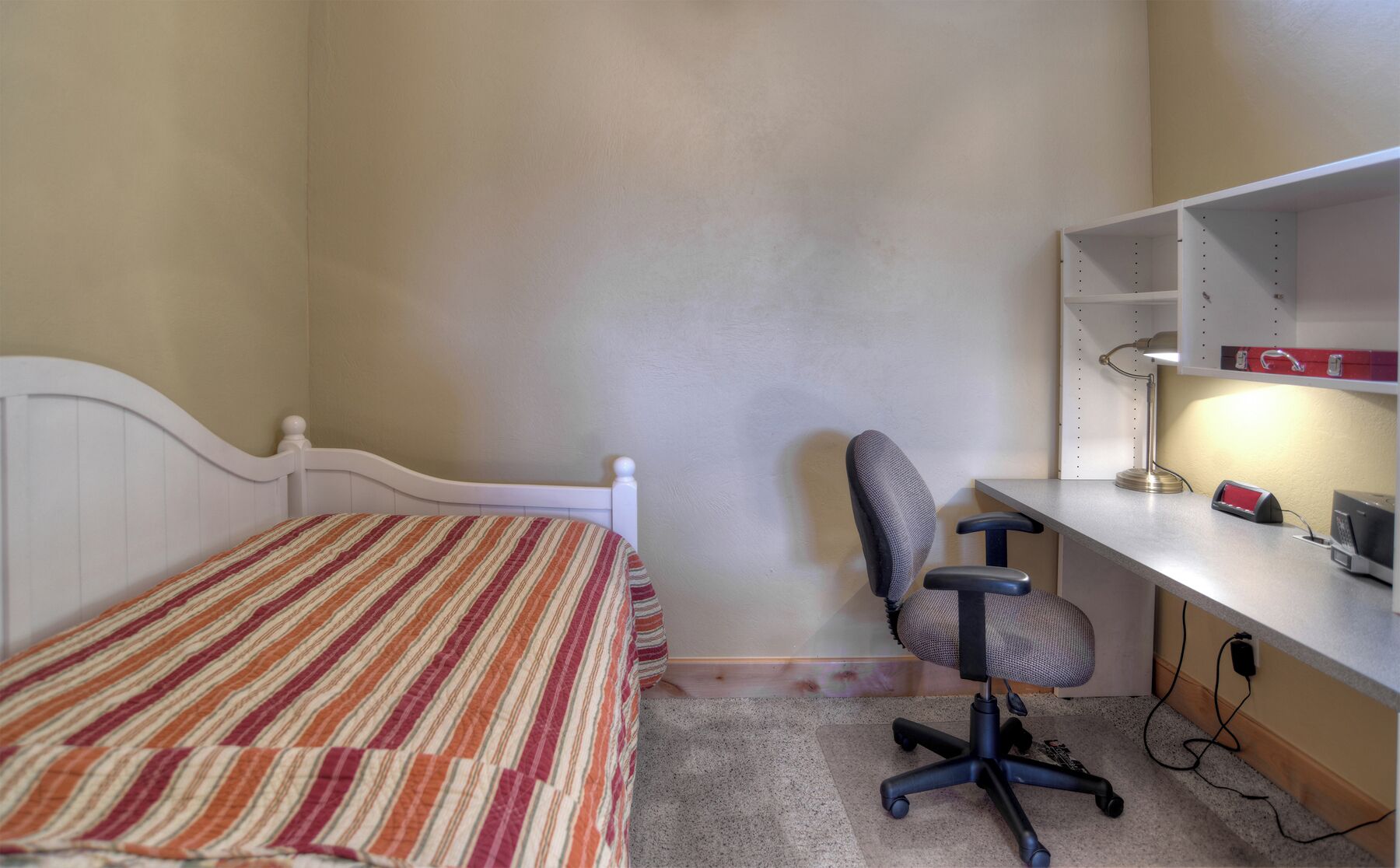 Een slaapkamer met een enkel bed, een bureau met stoel, en een plank met lamp.