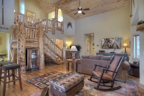 A spacious living room with a wooden staircase, a large flat-screen TV, and a cozy seating area with a patterned rug. - New! River Access Retreat | Golf Discount + 30 Min to Wolf Creek (South Fork)