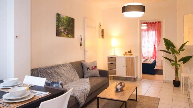 A modern living room with a sofa, coffee table, and dining area. There is a lamp, a plant, and a view into another room with a bed. - Apartment for 2/3 people - Ney District, Angers (Angers)